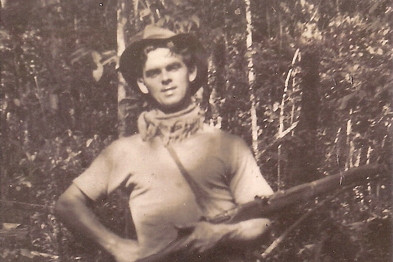 A black and white photograph of a man wearing a wide-brimmed hat, t-shirt and gun in a forest.