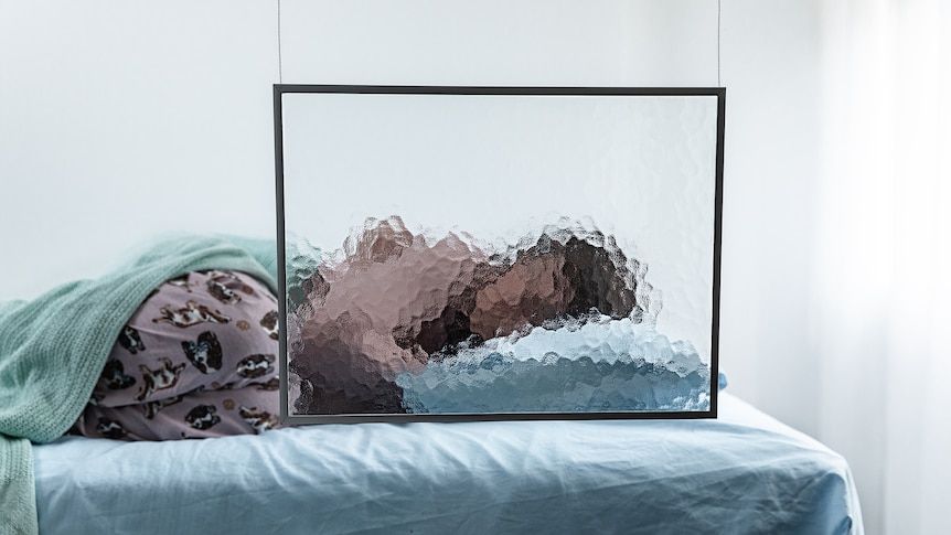 A girl curls up on a bed in a hospital. A frame of tempered glass blurs her face.