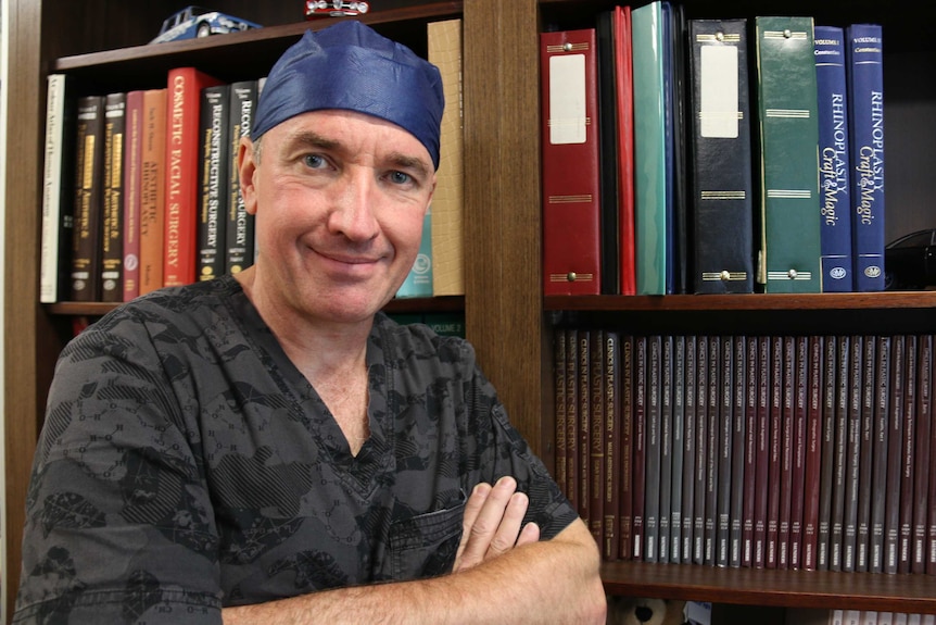 A man in scrubs stands in front of a book case with his arms folding.