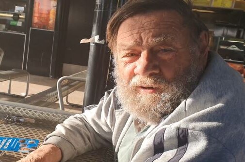 Een oudere, bebaarde man zit aan een tafel onder wat een patio blijkt te zijn.
