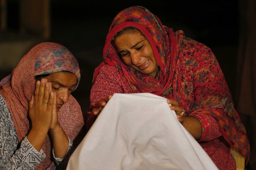 Two women wearing scarfs cry at the bottom of a body that is covered in white sheet