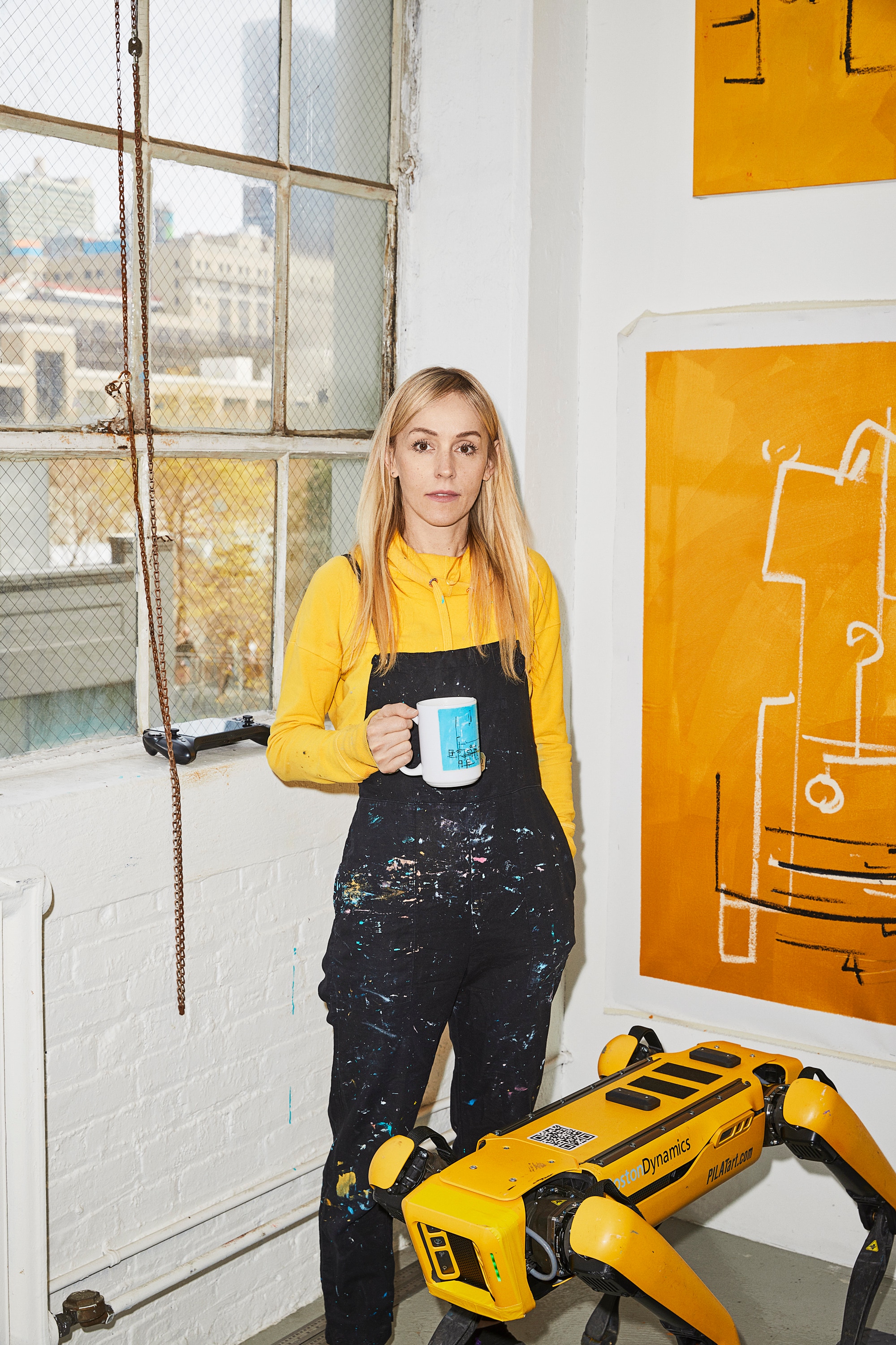 A middle-aged woman with long blonde hair stands in a studio space, wearing paint-covered overalls, with a yellow robot dog.