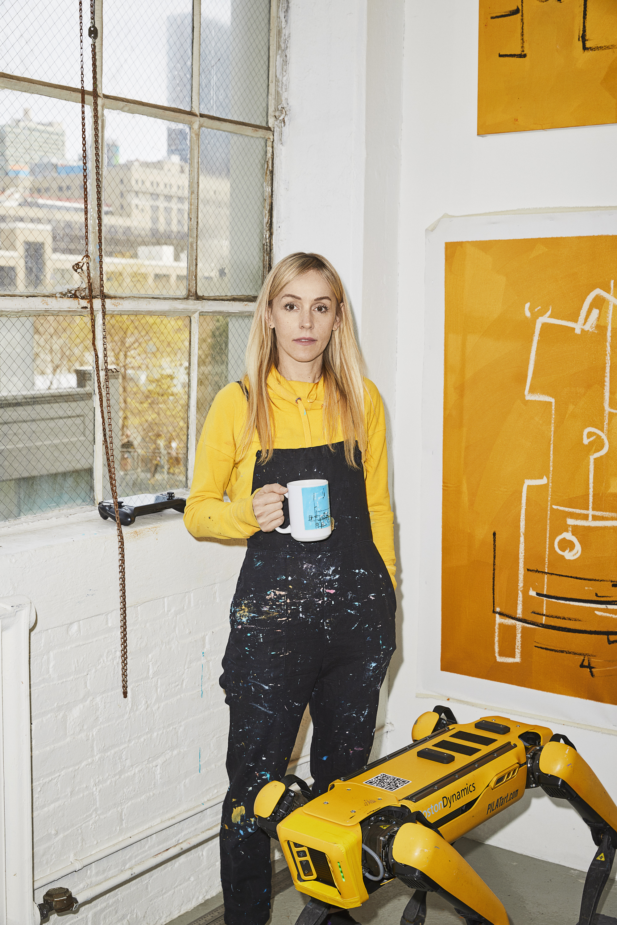 A middle-aged woman with long blonde hair stands in a studio space, wearing paint-covered overalls, with a yellow robot dog.