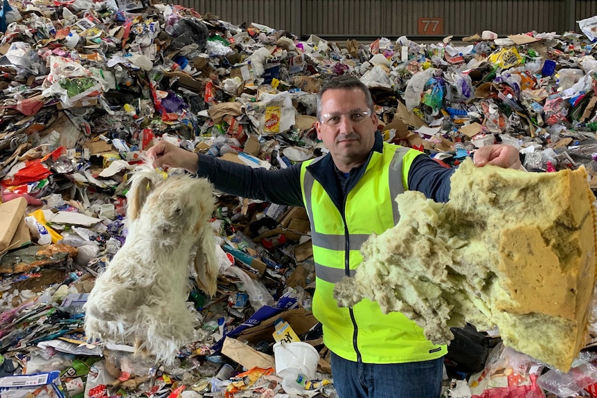 A man wearing a high-vis vest holds up foam insulation in front of a huge pile of waste to be recycled.