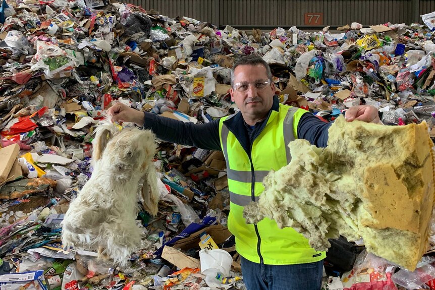 A man wearing a high-vis vest holds up foam insulation in front of a huge pile of waste to be recycled.