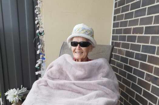 A smiling elderly woman in a warm hat and sunglasses sits under a rug on a balcony.