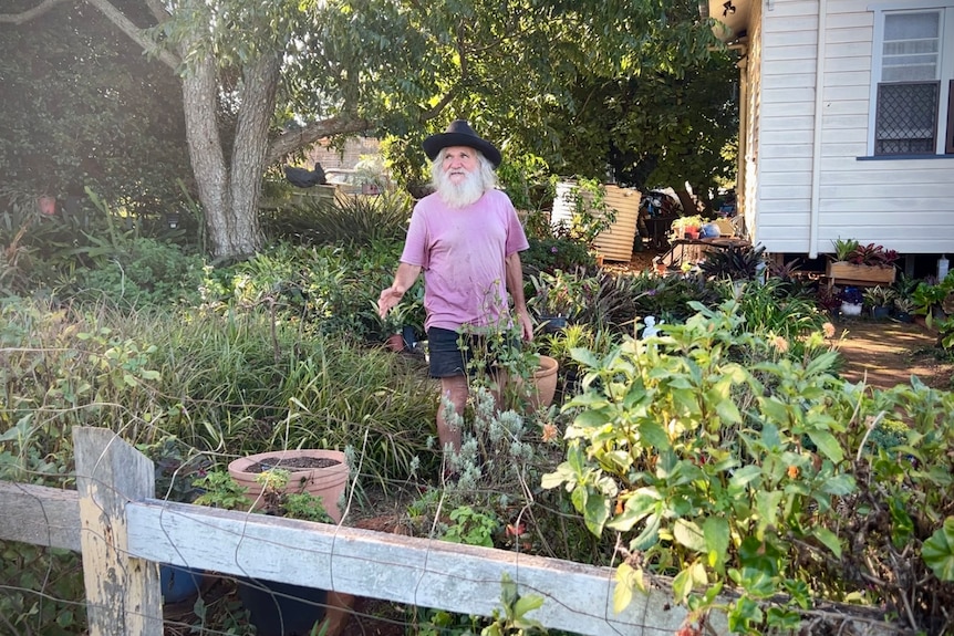 A man with a long grey beard and wearing a hat standing in a yard.