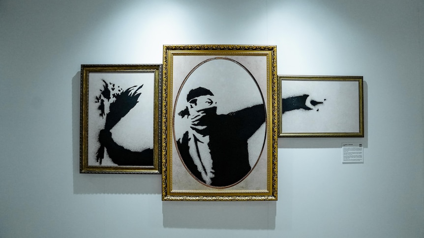 A Banksy mural of a man throwing a bunch of flowers is pictured across three gold frames.