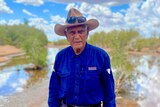 An elderly Indigenous man in a dark shirt and a hat, standing in front of a waterway fringed with trees. 