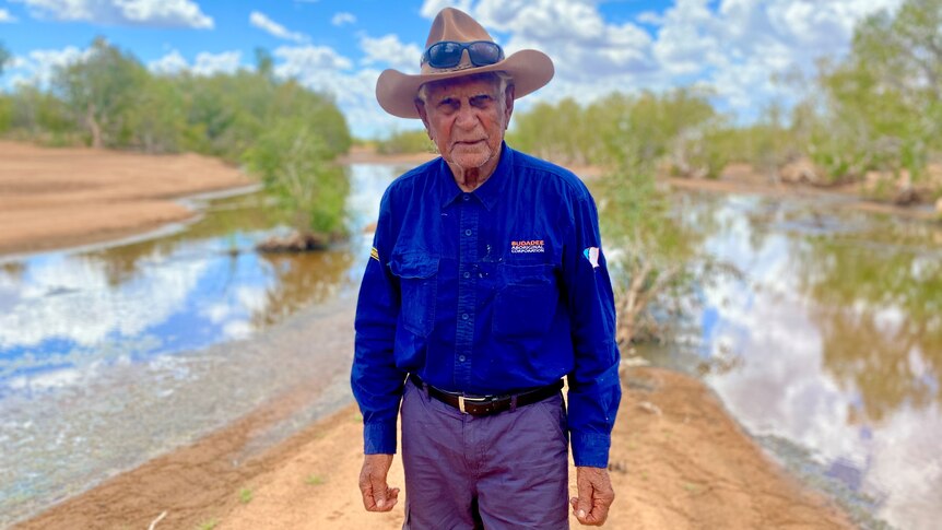 Stephen Stewart wears long blue shirt and hat with water and trees in the background. 