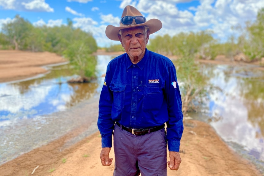 An elderly Indigenous man in a dark shirt and a hat, standing in front of a waterway fringed with trees. 