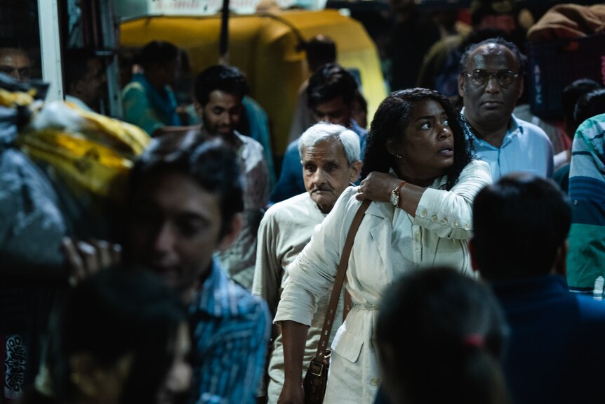 A middle-aged Black woman in a white dress walks through a busy crowd, looking alert.