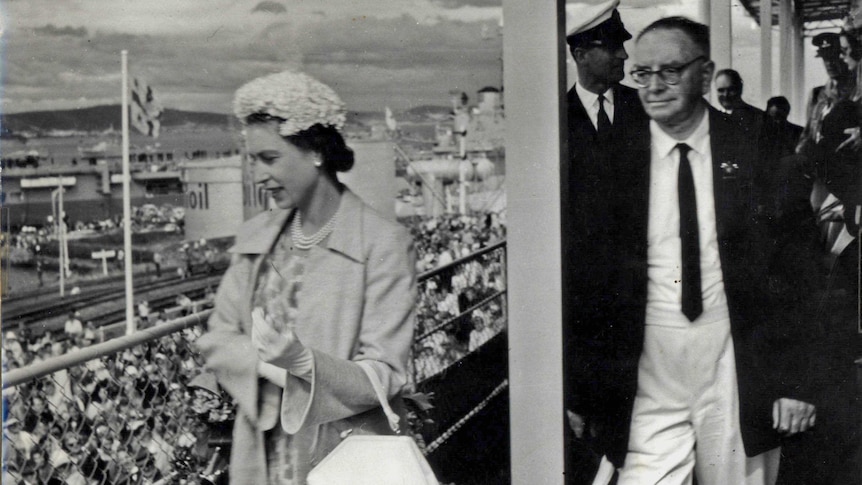 Picture of the Queen wearing pearls and a hat and white gloves and a man wearing a suit with crowds of people in the background