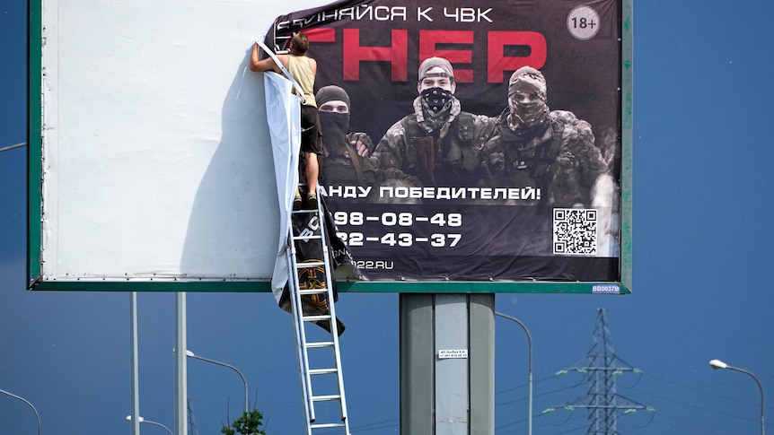 A man on a ladder pulls down the print of an enormous billboard ad showing men in military fatigues