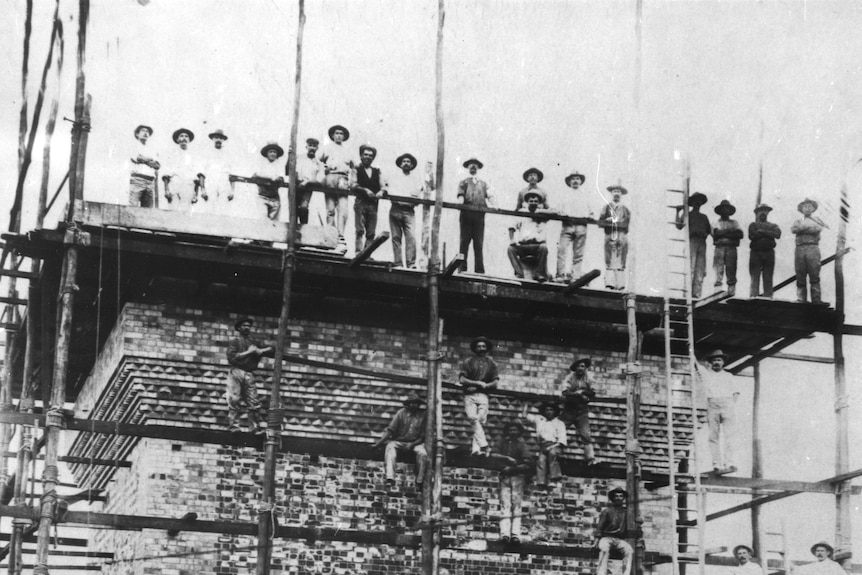 A black and white old photo of labourers standing on roof of brick building