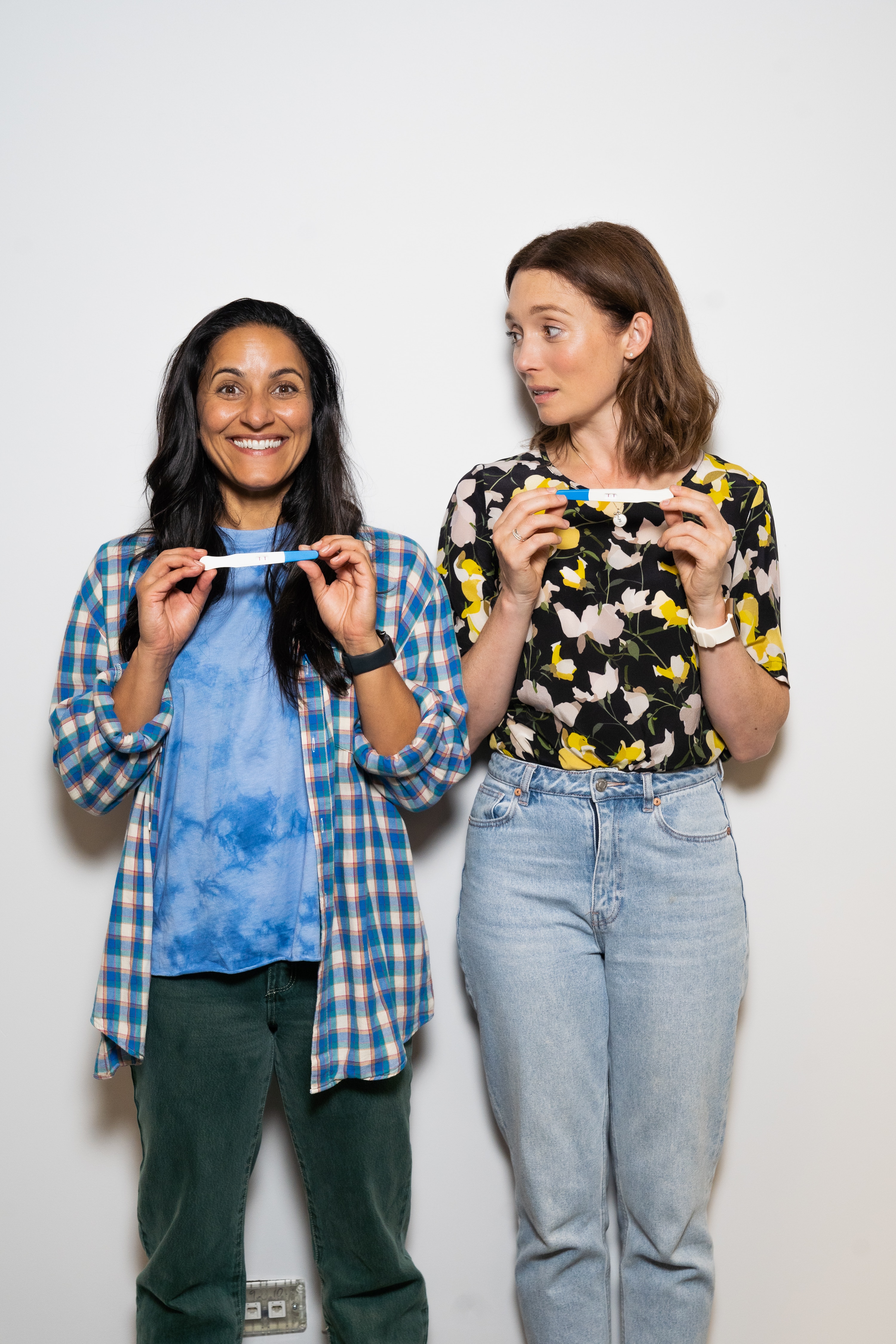 Two women stand next to each other holding pregnancy tests, the one on the left beaming and the one on the right shocked.