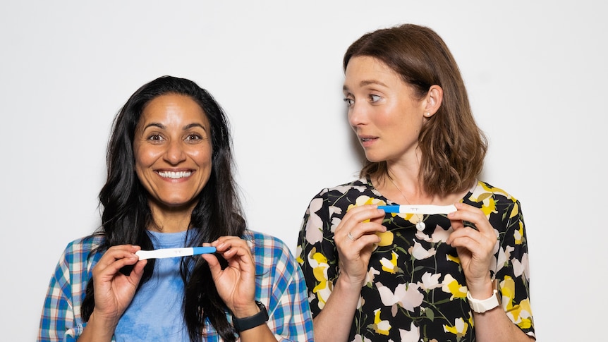 Two women stand next to each other holding pregnancy tests, the one on the left beaming and the one on the right shocked.