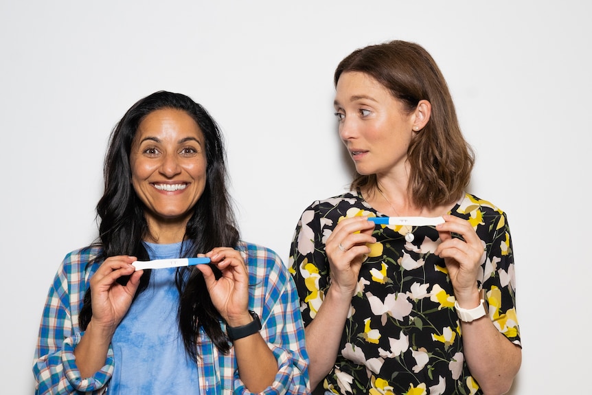 Two women stand next to each other holding pregnancy tests, the one on the left beaming and the one on the right shocked.