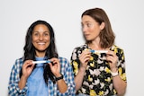Two women stand next to each other holding pregnancy tests, the one on the left beaming and the one on the right shocked.