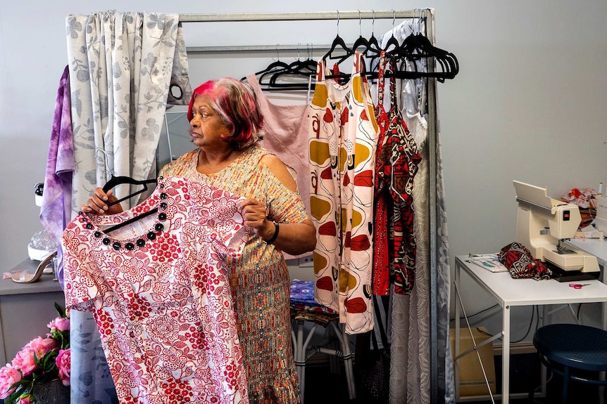 A woman stands with a clothing and a sewing machine sits in the corner.