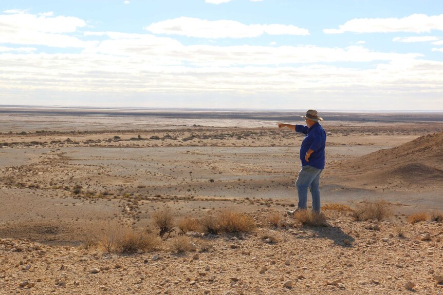 Wide shot of a man pointing away from camera across a vast, open outback landscape.