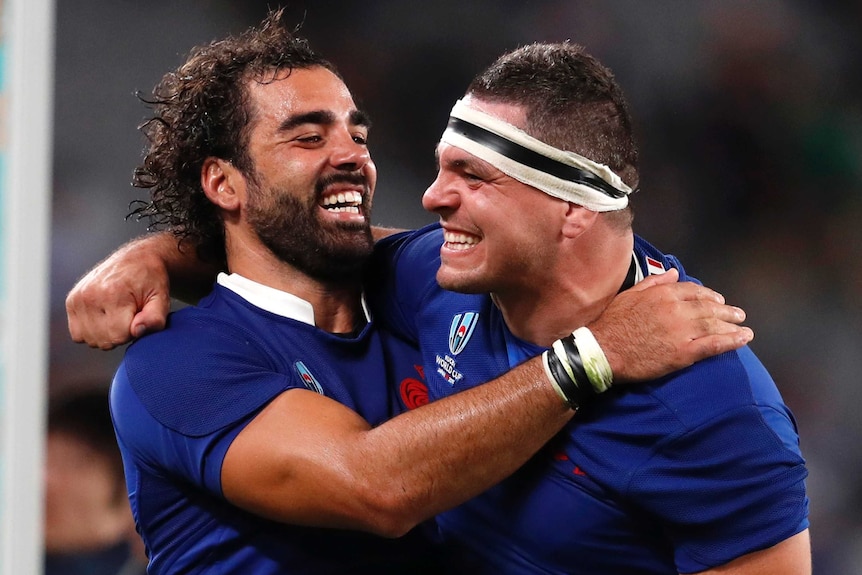 Two male rugby union players smile as they embrace to celebrating winning a match.