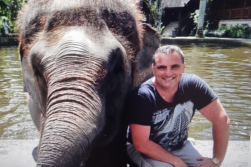 Peter kneels, smiling, in a t-shirt and shorts beside an elephant in front of a body of water.