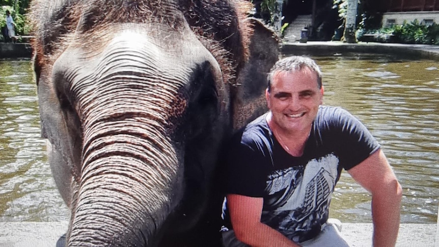 Peter kneels, smiling, in a t-shirt and shorts beside an elephant in front of a body of water.