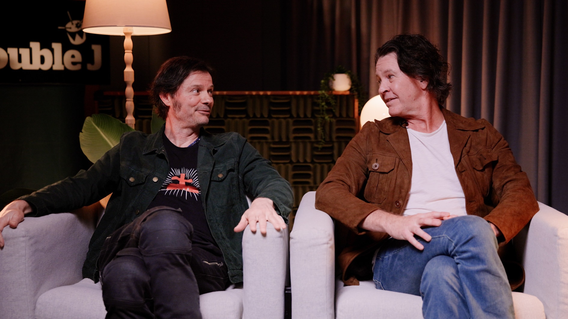 Darren Middleton and Bernard Fanning sit side by side in armchairs and look at each other.