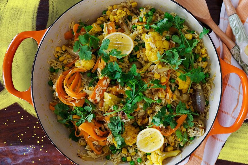 A cast iron pot with a cooked pilaf with cauliflower, carrot strips, corn, peas, topped with lemon and herbs.