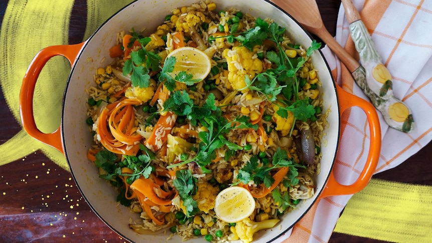 A cast iron pot with a cooked pilaf with cauliflower, carrot strips, corn, peas, topped with lemon and herbs, an easy recipe.