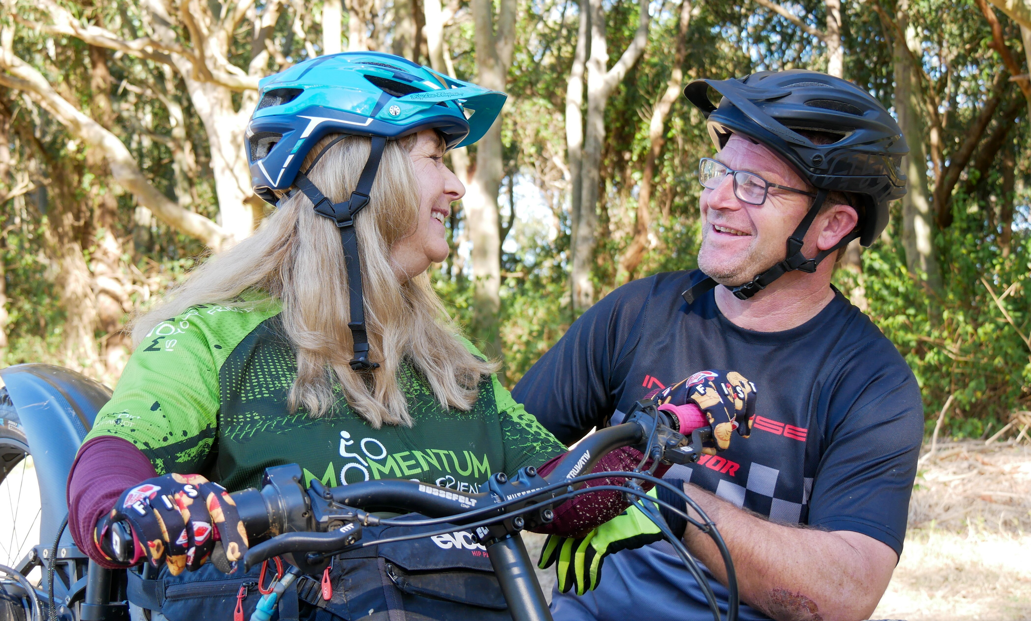 A woman sitting down on an adaptive bike wearing a helmet while smiling and looking at a man with a helmet and glasses.