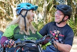 A woman sitting down on an adaptive bike wearing a helmet while smiling and looking at a man with a helmet and glasses.