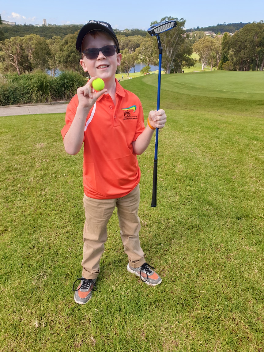 A nine year old boy dressed for golf smiles holding a golf ball in one hand and a golf club in the other
