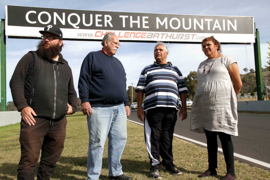 Four indigenous people stand in front of a sign which says conquer the mountain.