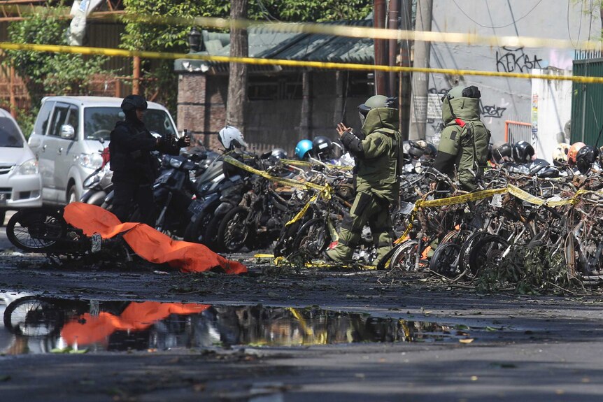 Two members of the bomb squad in protective suits stand near a stand of wrecked motorcycles, by a covered bike on its side