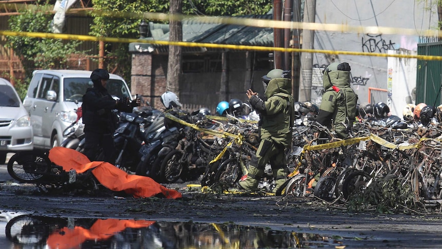 Two members of the bomb squad in protective suits stand near a stand of wrecked motorcycles, by a covered bike on its side