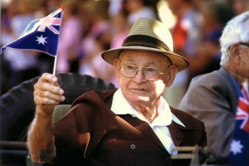 An old man wearing medals on his chest rides in a vehicle waving an Australian flag on an Anzac Day march.