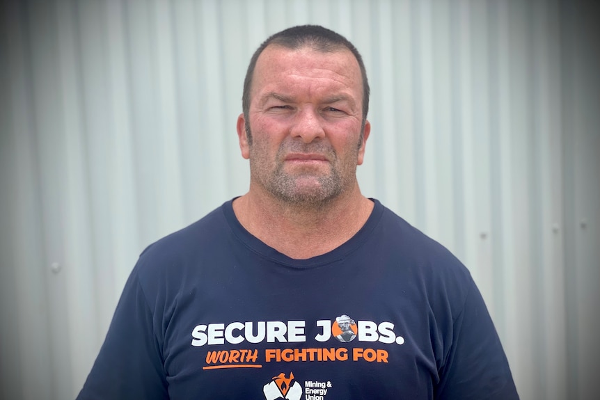 A man with closely cropped hair, wearing a blue t-shirt with secure jobs are worth fighting for, looks seriously at the camera. 