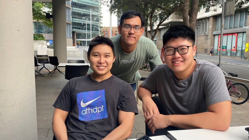 Three university students gather outside a cafe at a table. They smile for the camera.