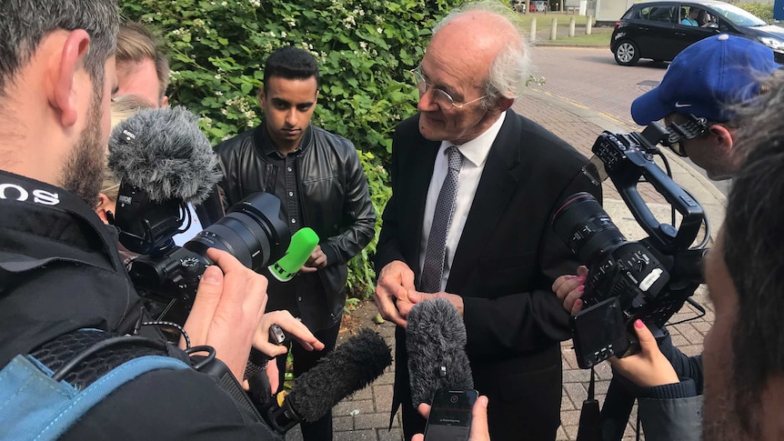 As elderly man wearing spectacles speaks to the media in the street.