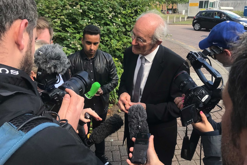 As elderly man wearing spectacles speaks to the media in the street.