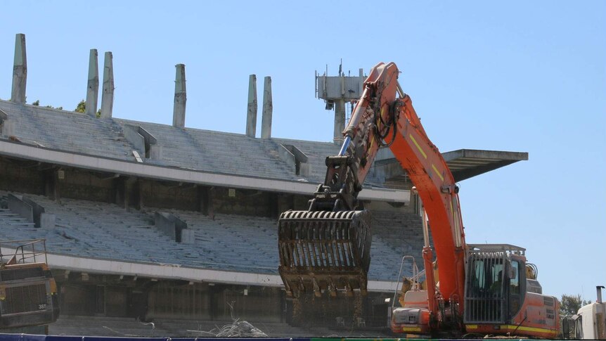 A construction vehicle picks up rubble with the remains of the old Subiaco Oval grandstand in the background.
