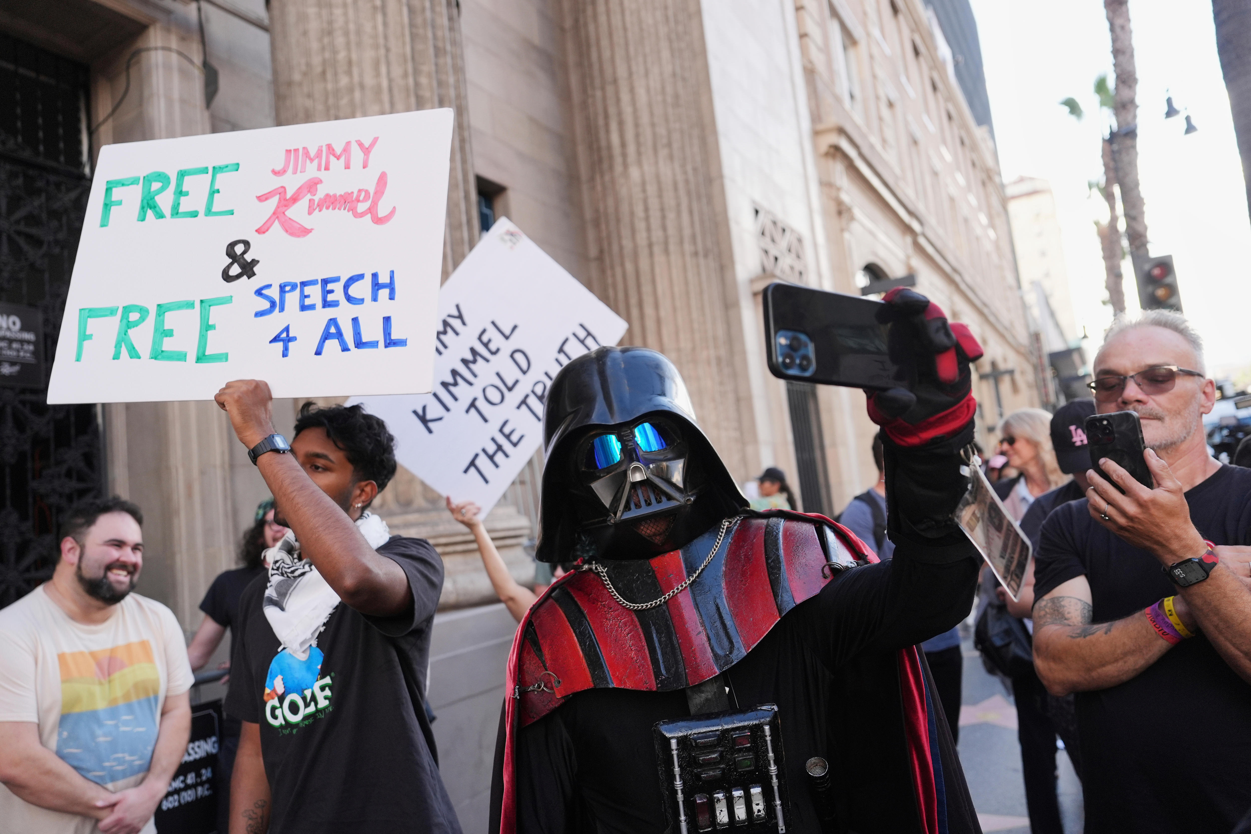 A person is dressed up as Darth Vader while protesters stand  behind. 