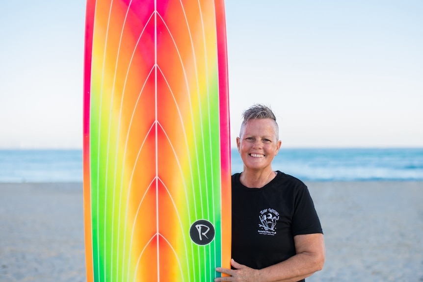 A woman standing beside a brightly coloured surfboard on a beach withe the sea behind her.