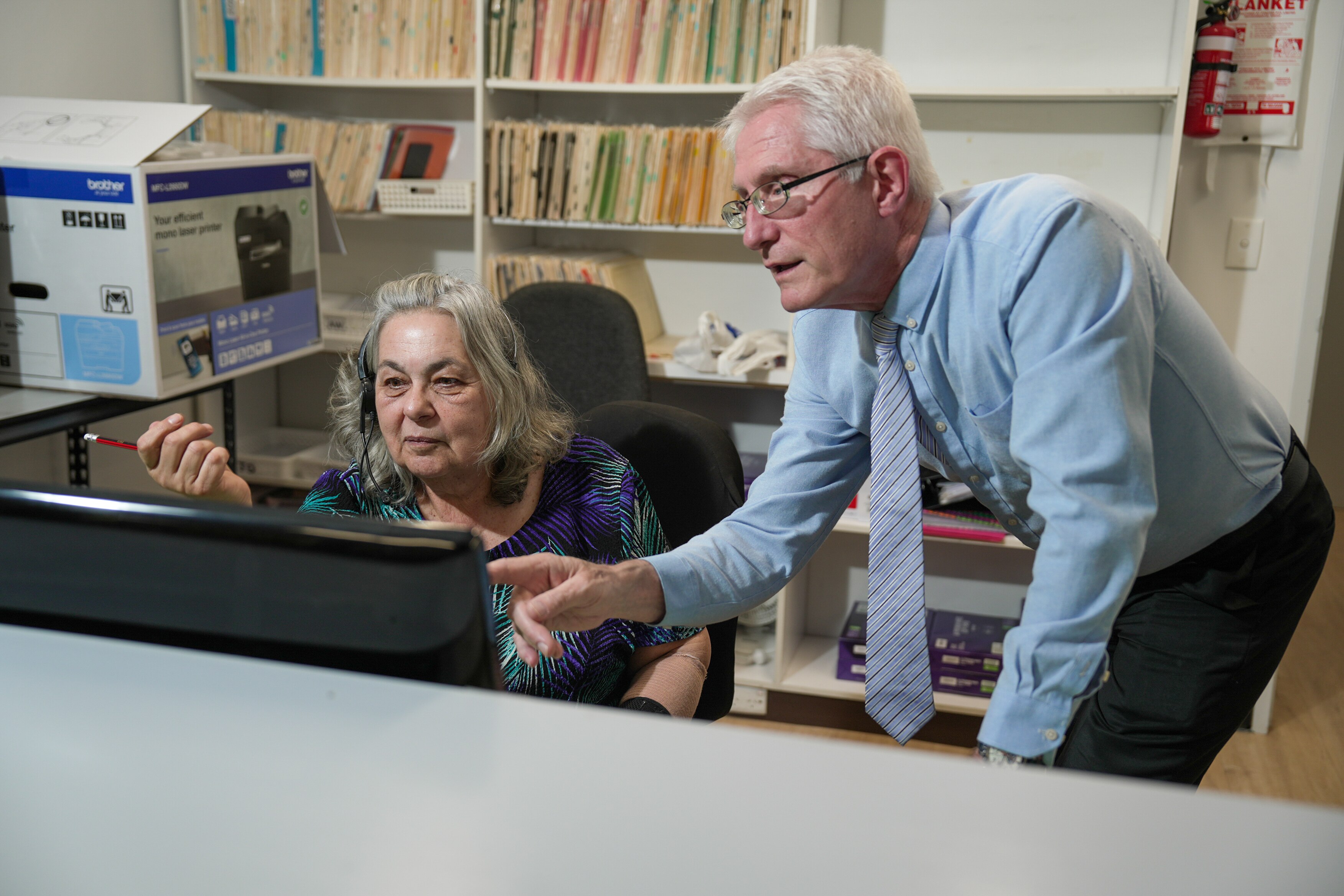 A woman sits at a computer behind a desk while a man stands next to her pointing at the screen
