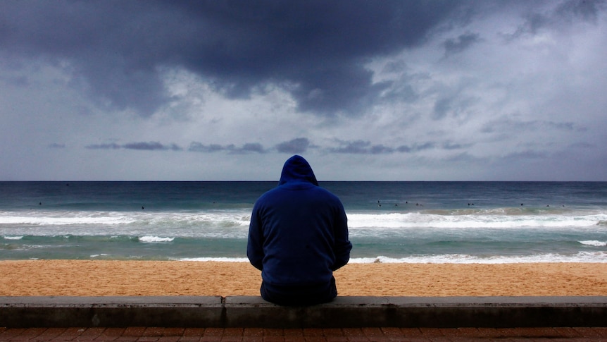 A man in a hoodie sits at a beach, looking out at the waves, as clouds gather