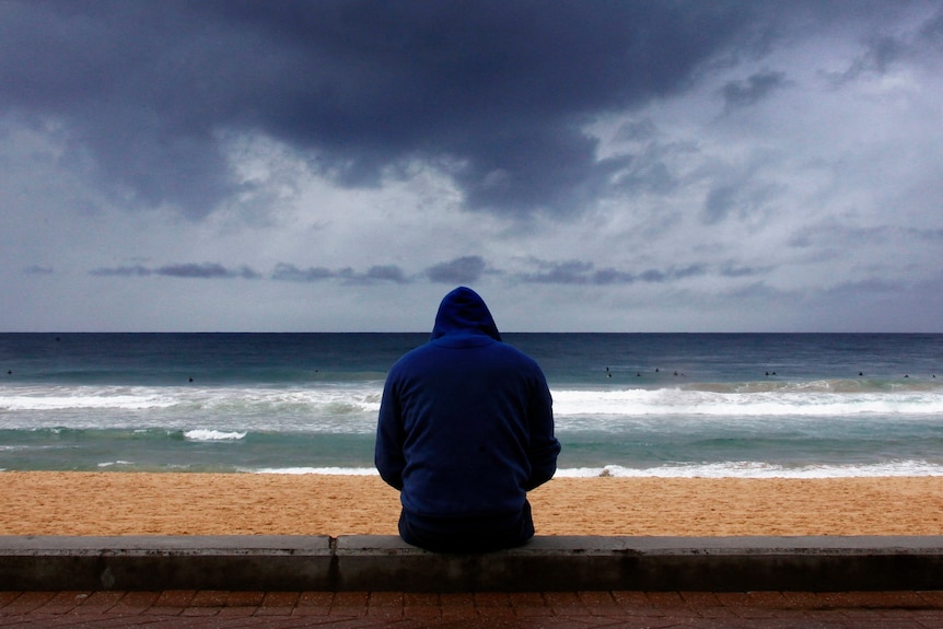 A man in a hoodie sits at a beach, looking out at the waves, as clouds gather