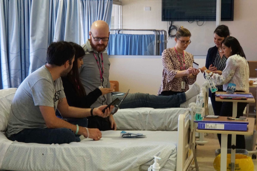 A team of medical researchers gather around hospital beds with folders and paperwork, speaking to trial participants.