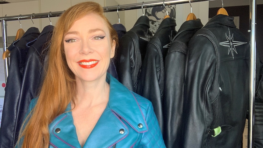 A woman with long red hair, wearing a bright blue leather jacket, stands in front of a rail of jackets.